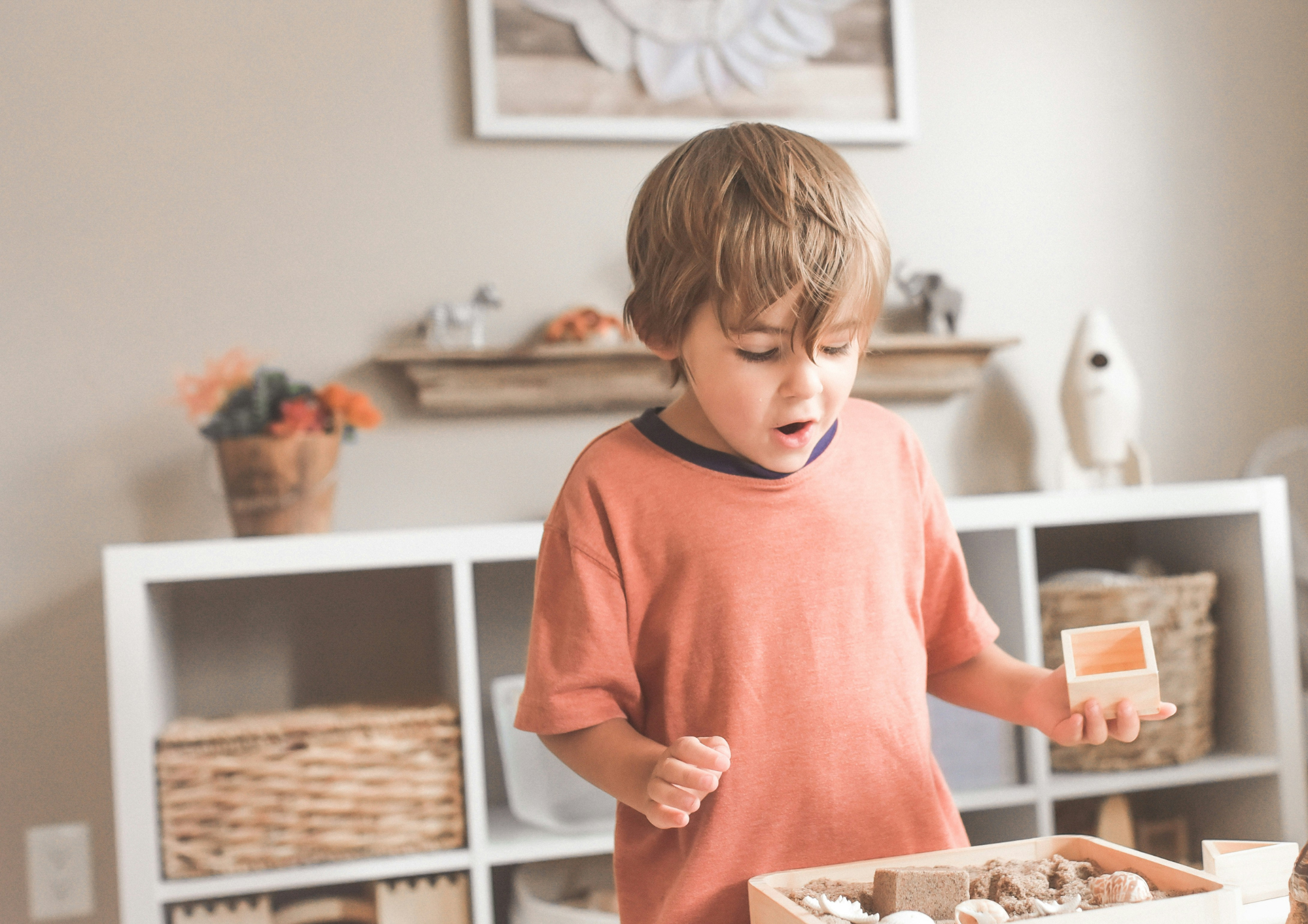A little boy is in a nicely organized playroom. He is playing at a table with a tray of open ended toys in front of him